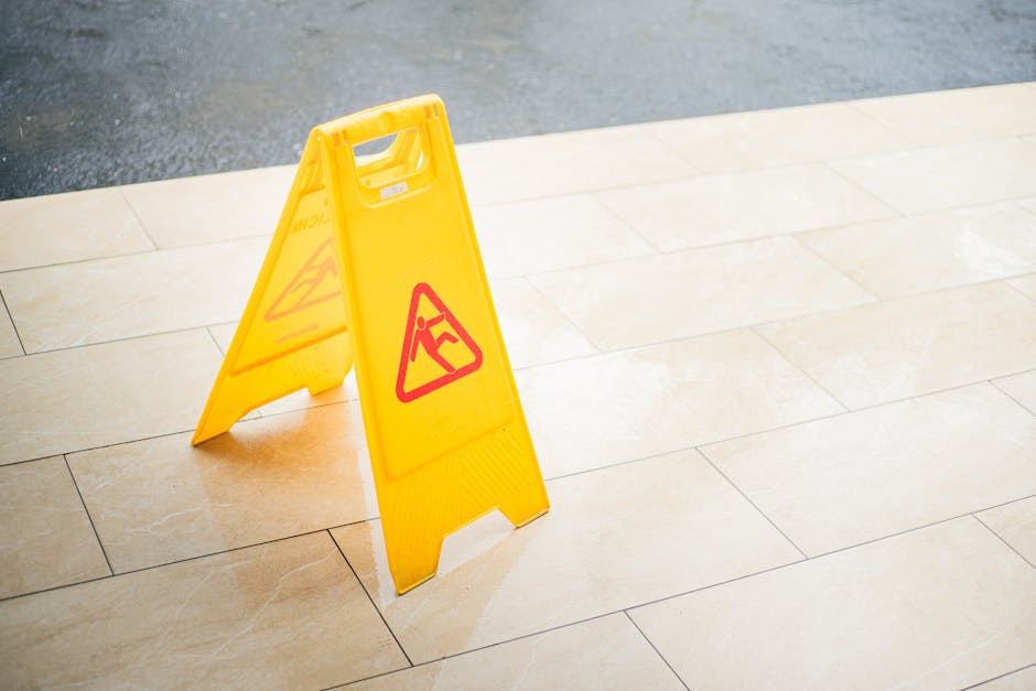 A yellow wet floor warning sign with black and red symbols stands on a beige tiled floor near an outdoor or indoor area, indicating a caution for slipping. The sign is made of plastic, with a foldable