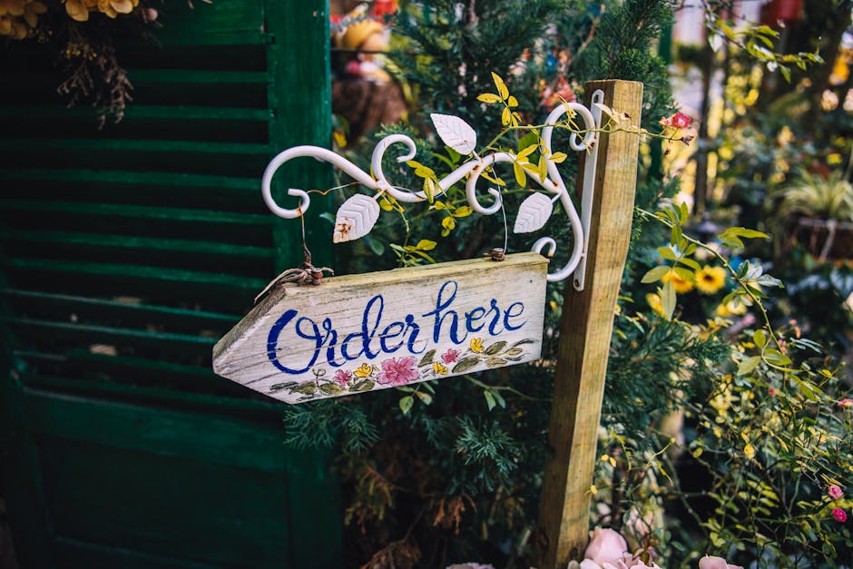 A rustic wooden garden sign with the words 'Order here' painted in blue, hanging from an ornate white metal bracket attached to a wooden post. Surrounding the sign are lush green foliage and flowering