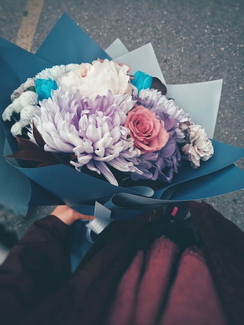 A vibrant floral bouquet featuring large white and lavender chrysanthemums, soft pink roses, and small blue accent flowers, all arranged in a lush, balanced manner. The bouquet is wrapped in elegant s