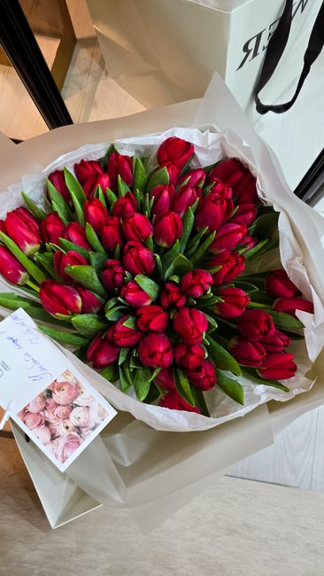 A large bouquet of fresh red tulips with green leaves arranged in a compact, rounded style. The tulips are wrapped in white tissue paper and placed inside a white gift box, positioned on a light-color