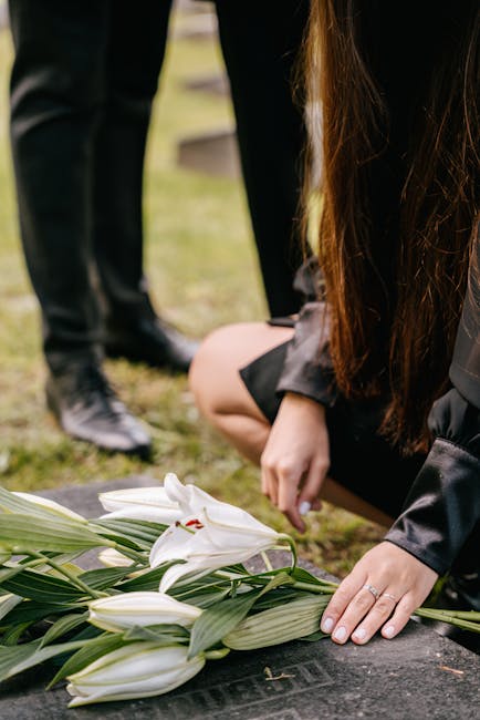 A person dressed in black is kneeling on the ground, placing a bouquet of white lilies with green stems and leaves on a flat surface, likely during a funeral or memorial service. The bouquet is wrappe