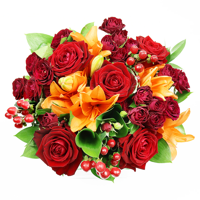 Captured directly overhead on a bright white surface, this circular bouquet reads as an intimate study in warm tones and plush textures. The composition is anchored by several large, fully bloomed deep red roses whose velvety petals curl outward to reveal layered centers; they convey classic romance and a tactile softness that draws the eye. Bright orange Asiatic lilies punctuate the design with trumpet-like forms-some flourishing wide with visible stamens, others poised in tight buds-introducing a kinetic contrast that suggests fresh arrival and unfolding beauty. Interspersed among these are dense clusters of small, deep burgundy spray roses, their compact heads offering a velvet-rich shadowed undertone that enhances the reds and balances the brighter oranges. Glossy Hypericum berries-rounded and jewel-like-appear in scattered bunches, adding reflective highlights and a playful, festive texture. Lush green foliage, probably ruscus or salal, peeks from between the blooms; its broad leaves provide a calming green frame and a clean, fresh counterpoint to the warm palette. The soft, even lighting emphasizes material qualities-satin lilies, velour roses, glassy berries and crisp leaves-without harsh shadows, making each element legible. This arrangement suggests occasions of deep feeling-anniversary surprises, heartfelt congratulations, or a centerpiece for a seasonal dinner-and often finds favour with customers across South Tottenham and nearby Lordship Recreation Ground gatherings who seek a bouquet that is both striking and tender.